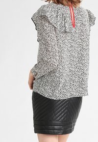 Blusa a pois bianca e nera con dettaglio a ruches sulle spalle, caratterizzata da un'apertura rossa sul retro. Abbinata a una gonna a tubino nera in pelle trapuntata.