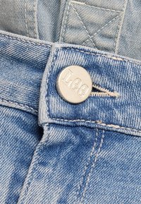 Ljusblå denimtyg har en metallknapp präglad med "Lee" och kontrasterande sömmar längs midjebandet.