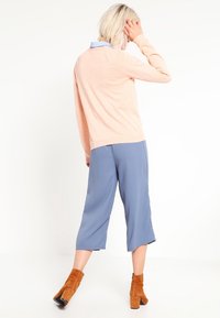 Un pull rose clair à manches longues, assorti à des culottes bleues amples et des bottes à cheville marron. Texture lisse et coupe décontractée.