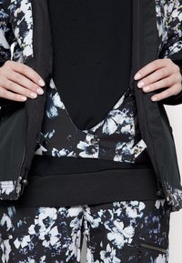 Händer som håller en öppen svart jacka med blommönstrad foder över matchande blommiga byxor och en svart skjorta med subtilt präglade bokstäver.