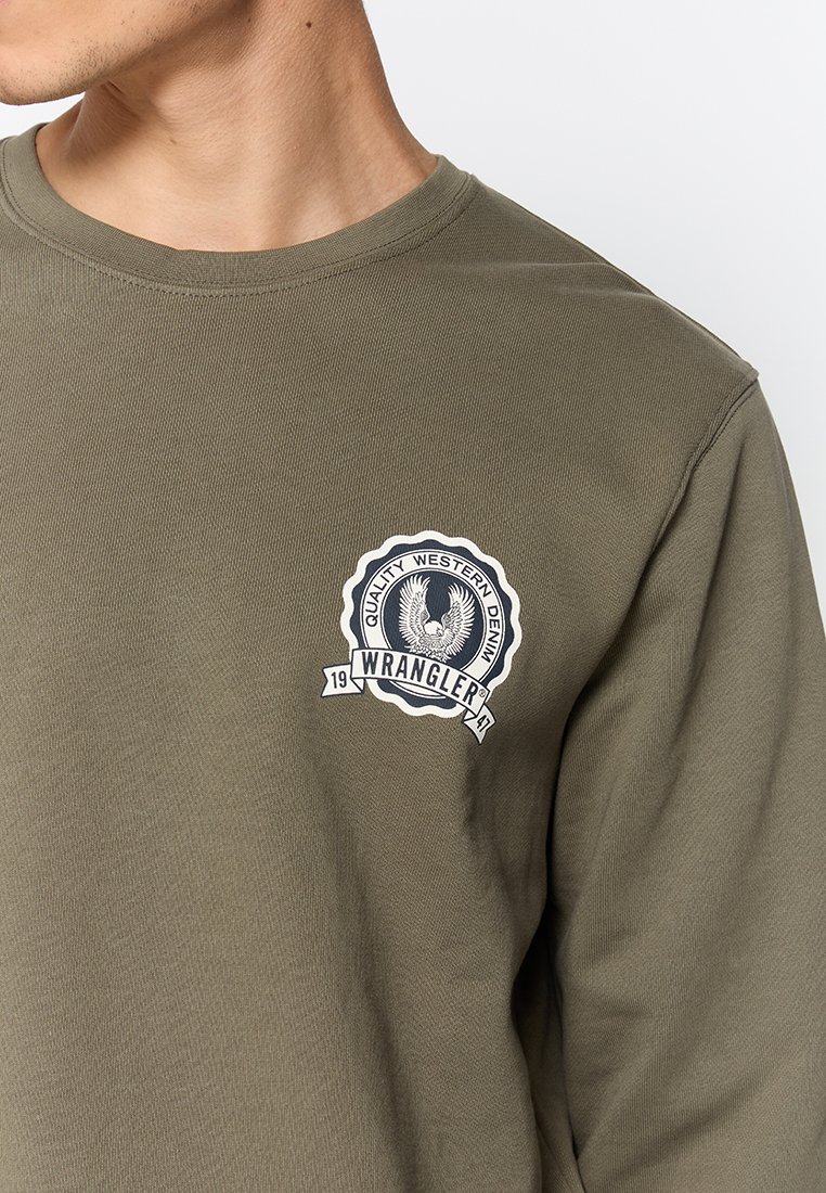 Oliwkowa koszula z długim rękawem wykonana z bawełny, z okrągłym dekoltem i okrągłą naszywką z logo w kolorze białym i niebieskim na klatce piersiowej.