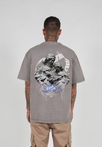 Grå oversized t-shirt med grafisk tryk af to skulpturelle figurer på ryggen, med blå accenter og lynmotiver.