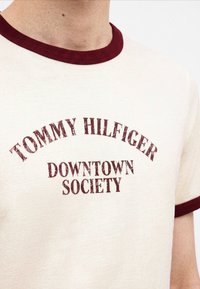 Μπεζ T-shirt με μπορντό γιακά και άκρα μανικιών, με μπορντό κείμενο "Tommy Hilfiger Downtown Society" στο στήθος.