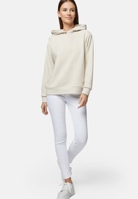 Jasno-beżowa bluza z kapturem, z ściągaczami przy rękawach i u dołu, noszona na białych rurkach i białych sneakersach, stojąca na neutralnym tle.