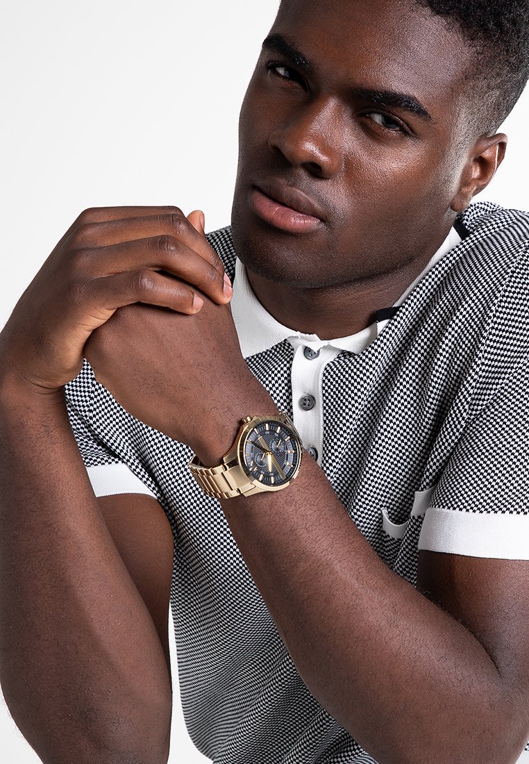 Camisa de polo a cuadros en blanco y negro, con cuello blanco, reloj de pulsera dorado con esfera azul, modelo posando con las manos entrelazadas.