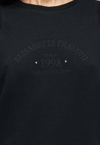 Μαύρο ύφασμα με ανάγλυφο κείμενο "Elisabetta Franchi Since 1998 Limited Edition" και δύο μικρές χρυσές καρφίτσες κοντά στο κέντρο του κειμένου.