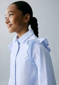 Vaaleansininen raidallinen paita, jossa on rypytetty yksityiskohta olkapäillä, napitettava kaulus ja pitkät hihat, joissa on sileä rakenne.