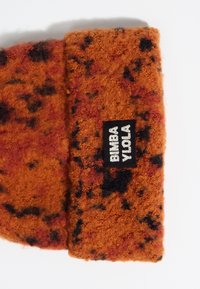 Tejido texturizado naranja con manchas negras y rojas. Presenta una etiqueta negra con "BIMBA Y SOLA" en texto blanco.