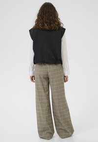 Göndör barna hajú személy fekete ujjatlan mellényben, fehér hosszú ujjú ingben és bézs kockás, széles szárú nadrágban áll, háttal a néző felé.