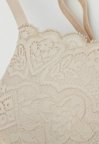 Beige tyg-bustier med intrikata hjärt- och blommönster i spets, texturerad yta och vågiga kanter med mjuk rem.