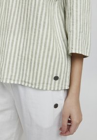 Top de algodón a rayas en verde claro y blanco, con mangas tres cuartos y detalles en negro en el dobladillo, combinado con pantalones blancos.