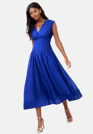 Vrouw die een koningsblauwe geplooide midi-jurk draagt met een V-hals en korte mouwen, gecombineerd met witte sandalen met hoge hakken.