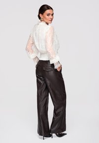 Femme en blouse blanche en dentelle transparente et pantalon large en cuir noir, posant les mains dans les poches, regardant par-dessus l'épaule.