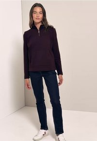 Sweat-shirt bordeaux zippé avec une poche avant, associé à un jean foncé et des baskets blanches avec des accents rouges. Tissu doux et décontracté.