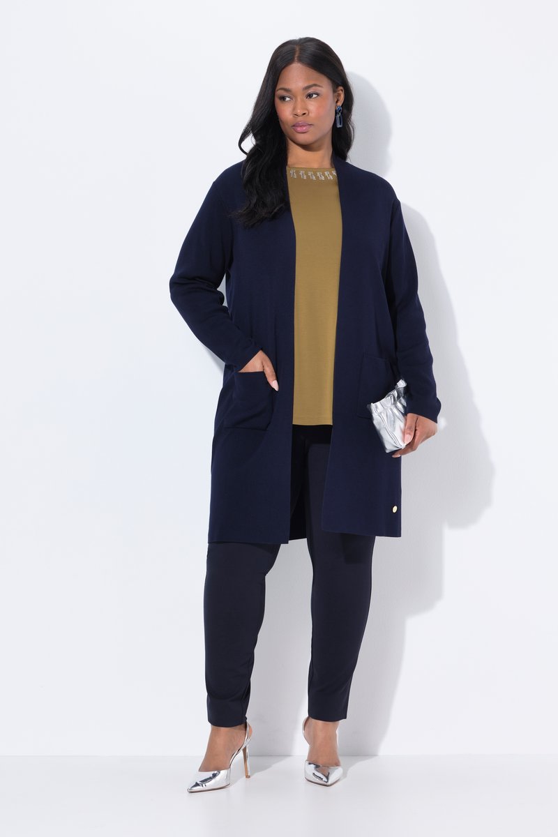 Mujer de pie con cárdigan azul marino, camiseta oliva, pantalones azul marino, tacones altos plateados, sosteniendo un bolso de mano plateado, con fondo blanco liso.