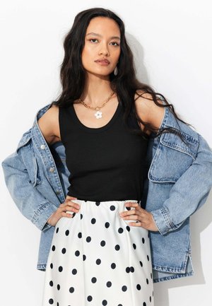 Femme aux longs cheveux foncés portant un débardeur noir, une jupe blanche à pois noirs, une veste en jean tombant des épaules et une chaîne en or avec un pendentif fleur.