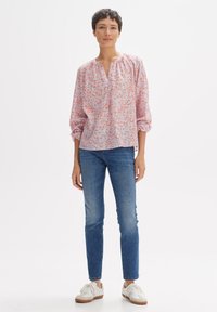 Blusa a maniche lunghe rosa chiaro con motivo floreale blu e corallo, caratterizzata da un collo rotondo e maniche arricciate. Abbinata a jeans blu e sneakers bianche.