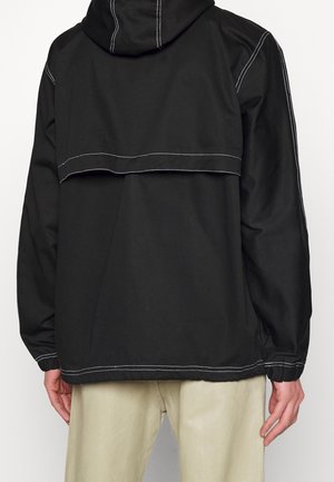 Rückansicht einer Person, die eine schwarze Kapuzenjacke mit weißen Nähten und beige Hosen trägt und vor einem einfarbigen Hintergrund steht.