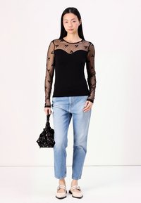 Top negro de manga larga con tela de malla transparente que presenta un patrón de puntos y acentos florales, combinado con jeans azul claro y un bolso negro.