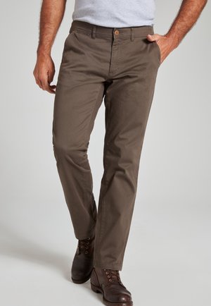 Homme portant un pantalon marron coupe droite, une chemise gris clair et des bottes en cuir marron foncé, debout avec une main dans la poche.