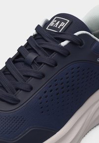 Chaussure de sport bleu marine avec tige en mesh perforé, lacets bleus marins, semelle blanche et logo GAP sur la languette et le côté.