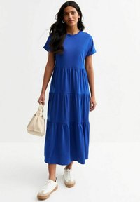 Blue, short-sleeved, tiered maxi dress made of soft fabric. Worn with white sneakers and a beige handbag.
