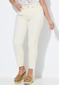 Béžové džíny s květinovým laser-cut vzorem po straně, s úzkým střihem a pěti kapsami. Kombinované s hnědými sandály na klínu.