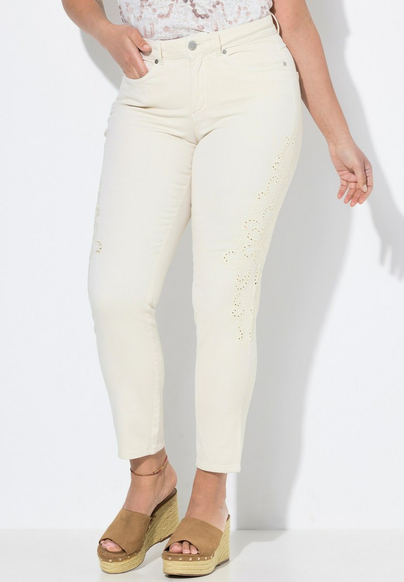 Béžové džíny s květinovým laser-cut vzorem po straně, s úzkým střihem a pěti kapsami. Kombinované s hnědými sandály na klínu.
