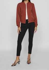 Rostfarbene Bomberjacke mit Reißverschluss, gerippten Bündchen und zwei seitlichen Reißverschlusstaschen, kombiniert mit schwarzen Skinny-Jeans und Riemchenabsätzen.