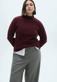 Burgunderfarbener Rippenpullover mit hohem Kragen, kombiniert mit hellgrauen, maßgeschneiderten Hosen über einem weißen Hemd. Glatte Textur, lässige Passform.