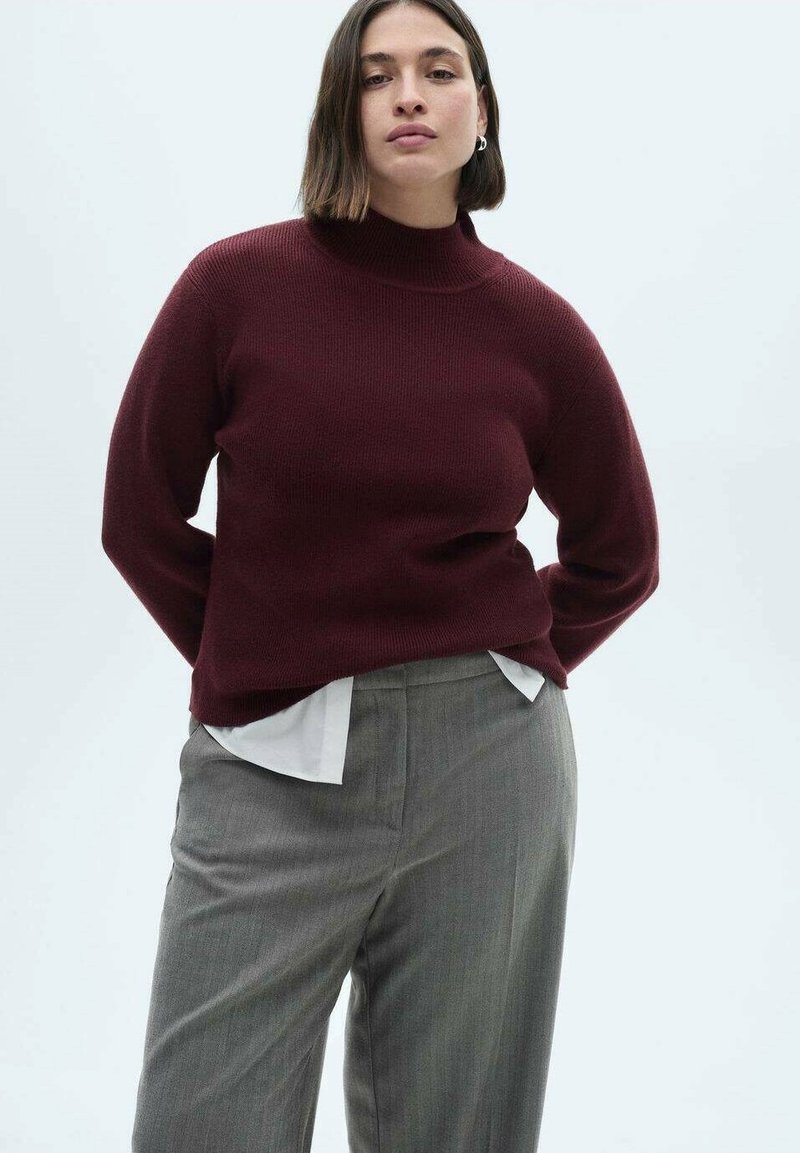 Burgunderfarbener Rippenpullover mit hohem Kragen, kombiniert mit hellgrauen, maßgeschneiderten Hosen über einem weißen Hemd. Glatte Textur, lässige Passform.