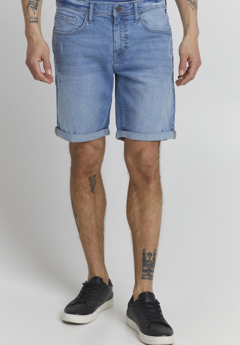 Lyse blå denimshorts med oppbrettede ermer, fem lommer og lett slitasje. Båret med svarte joggesko, som viser frem eksponerte tatoveringer på bena.