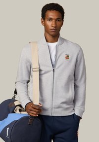 Mladý muž v svetlosivej bunde na zips a námorníckych nohaviciach drží námornícku a svetlomodrú športovú tašku s béžovým popruhom cez rameno.