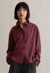 Burgundy button-up shirt with a relaxed fit, long sleeves, and a collar. Smooth texture, subtly iridescent finish. Paired with dark trousers.