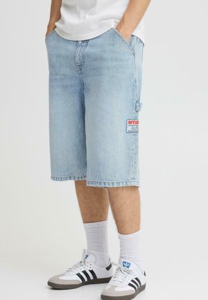 Lys blå denimshorts med en avslappet passform, med en sidelomme og en "BITUM"-etikett på høyre lår. Brukes med hvite sokker og joggesko.