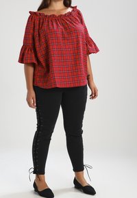 Haut à carreaux rouges hors des épaules avec des manches volantes, associé à un jean skinny noir avec des détails à lacets sur les côtés. Des ballerines noires complètent le look.