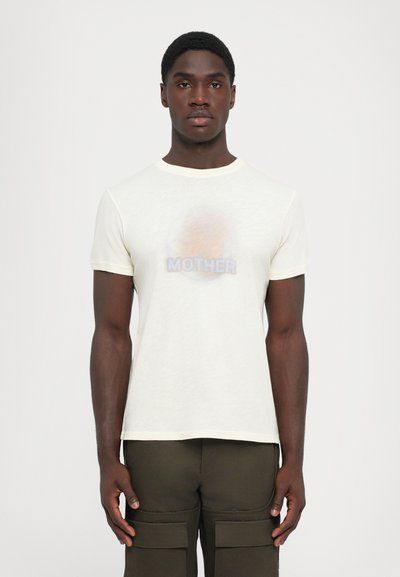 Young man with dark skin wearing a translucent white T-shirt with "MOTHER" text, paired with dark green cargo pants, standing against a plain white background.