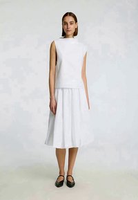 Donna in piedi che indossa una maglietta senza maniche bianca, una gonna midi bianca e delle scarpe Mary Jane nere, su uno sfondo chiaro e semplice.