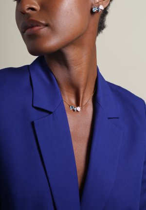 Gouden ketting met drie gekleurde edelsteen hangers—blauw, roze en helder—gecombineerd met een koningsblauwe blazer, die een gladde stofstructuur toont.