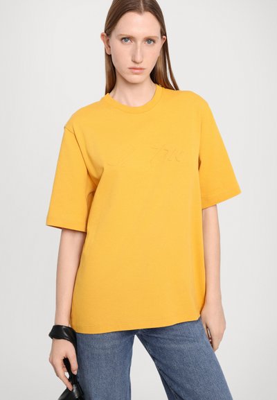 Kollane puuvillane T-särk lühikeste varrukatega ja ümmarguse kaelusega, millel on reljeefne logo detail. Kandmine koos siniste teksadega.