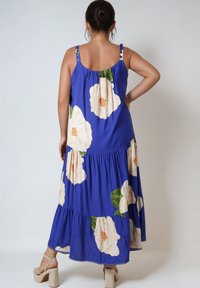 Maxi-jurk van blauwe stof versierd met grote crèmekleurige en groene bloemenpatronen, met verstelbare bandjes met knopen en een gelaagd ontwerp.
