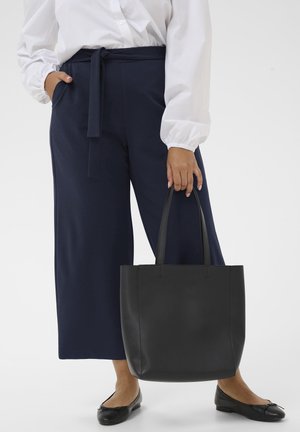 Osoba v bielej blúzke a tmavomodrých nohaviciach drží veľkú čiernu taška, stojí v čiernych baletných balerínach na jednofarebnom pozadí.