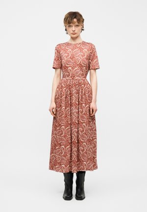 Žena stojaca, oblečená v kovovočervenom vzorovanom midi šatách s krátkymi rukávmi a čiernymi členkovými čižmami na bielom pozadí.