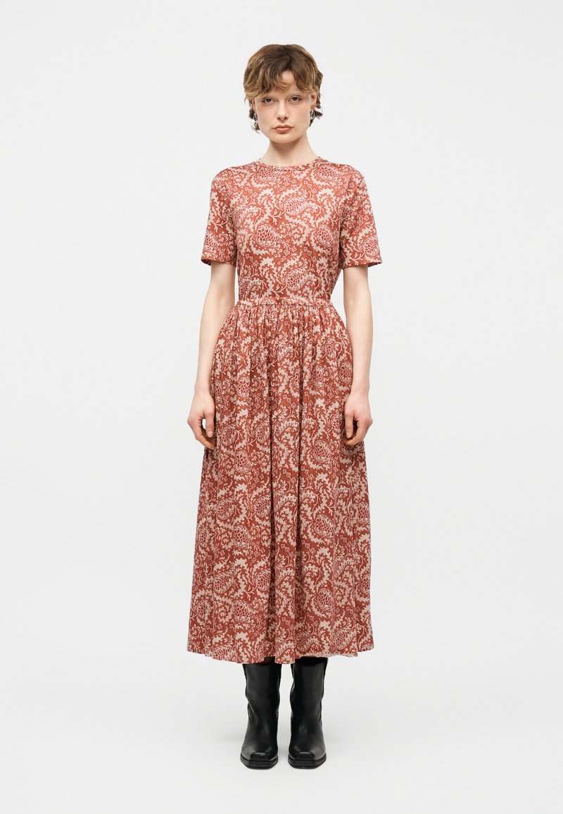 Γυναίκα που στέκεται φορώντας ένα μεσομήκες φόρεμα με μοτίβο σε σκουρόκοκκινο χρώμα, κοντά μανίκια και μαύρα μποτάκια μέχρι τους αστραγάλους, σε λευκό φόντο.