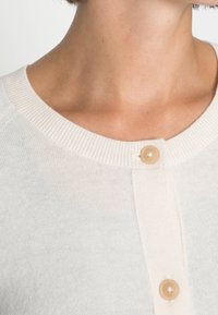 Ljusbeige cardigan med ribbad rund halsringning, enstaka beige knäppning och finstickad textur. Enkel och minimalistisk design.