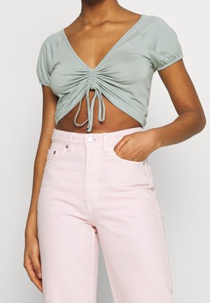 Frau trägt ein hellblau gerafftes cropped Top mit Kordeln und hoch taillierte hellrosa Hose, Hand in der Tasche.