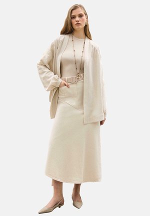 Žena v béžovej dlhej sukni, zladenom topu, vo veľkej bunde, s opaskom, dlhým náhrdelníkom a špicatými topánkami so zapínaním na pätu, stojaca s jednou rukou v vrecku.