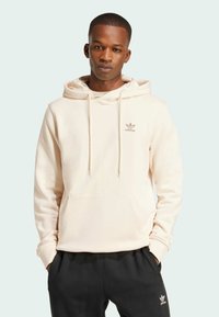 Light beige hoodie with a front pocket, drawstring hood, and small brown Adidas logo on the chest. Paired with black sweatpants.
