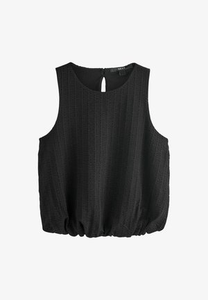 Femme portant un haut noir sans manches, une mini-jupe en denim foncé avec une fermeture éclair à l'avant et des sandales à boucle colorée, debout devant un fond uni.