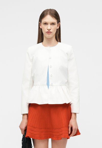 Γυναίκα που φοράει λευκό σακάκι με κουμπιά και πεπλουμ πάνω από μπλε τοπ και πορτοκαλί πλισέ μίνι φούστα, κρατώντας μαύρη τσάντα με κρόσσια.